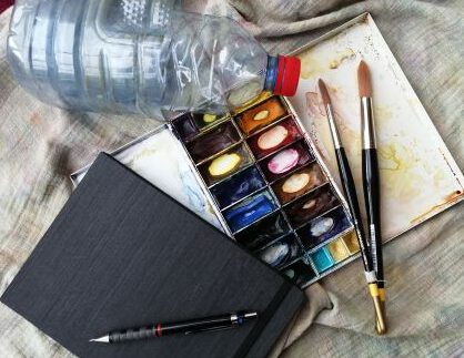 Das Foto zeigt ein Skizzenbuch mit Aquarellfarbkasten, Pinsel, Bleistift, Wassenflasche und Mallappen